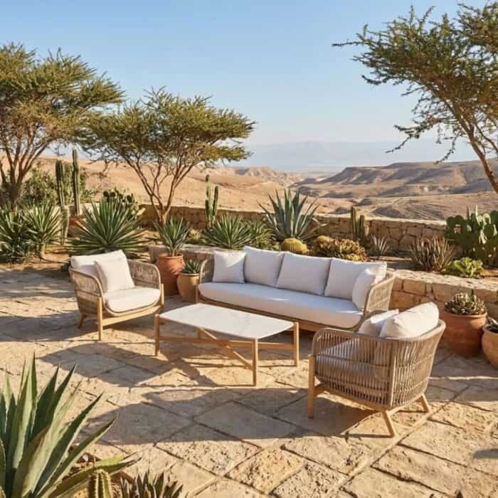 מרפסת חיצונית עם פינות ישיבה לגינה, הכוללת ספה, שני כיסאות גינה ושולחנות גינה המונחים על אריחי אבן, מוקפת בצמחייה ועצים מדבריים, המשקיפה על נוף גבעותי ויבש תחת שמיים בהירים.